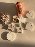 Suspensions de Noël artisanales en béton blanc avec incrustations de feuilles dorées, formes étoile, cœur et médaillon rond ondulé. Décorations faites main présentées avec perles décoratives et fil doré dans une ambiance lumineuse et élégante.