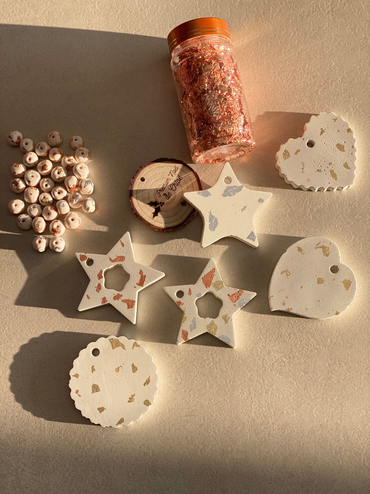 Suspensions de Noël artisanales en béton blanc avec incrustations de feuilles dorées, formes étoile, cœur et médaillon rond ondulé. Décorations faites main présentées avec perles décoratives et fil doré dans une ambiance lumineuse et élégante.