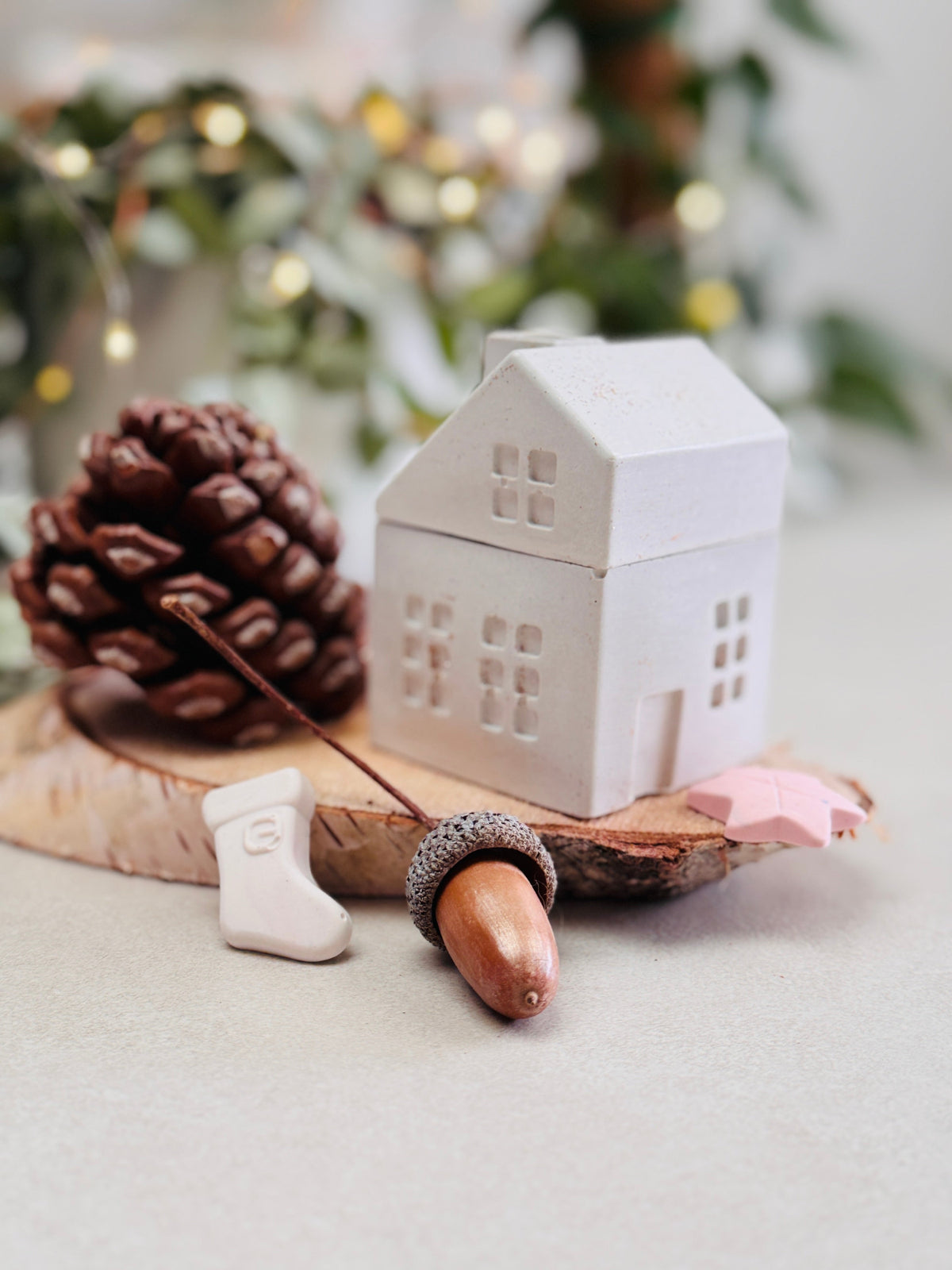 Bougie blanche en forme de maison avec cheminée posée sur un rondin de bois, entourée de petites décorations de Noël, de pommes de pin et de glands, lumières de Noël.
