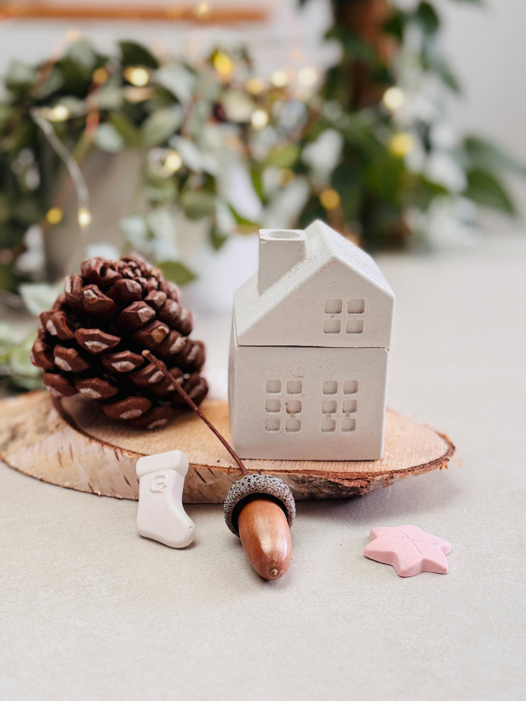 Bougie blanche en forme de maison avec cheminée posée sur un rondin de bois, entourée de petites décorations de Noël, de pommes de pin et de glands, chausson de père Noël.
