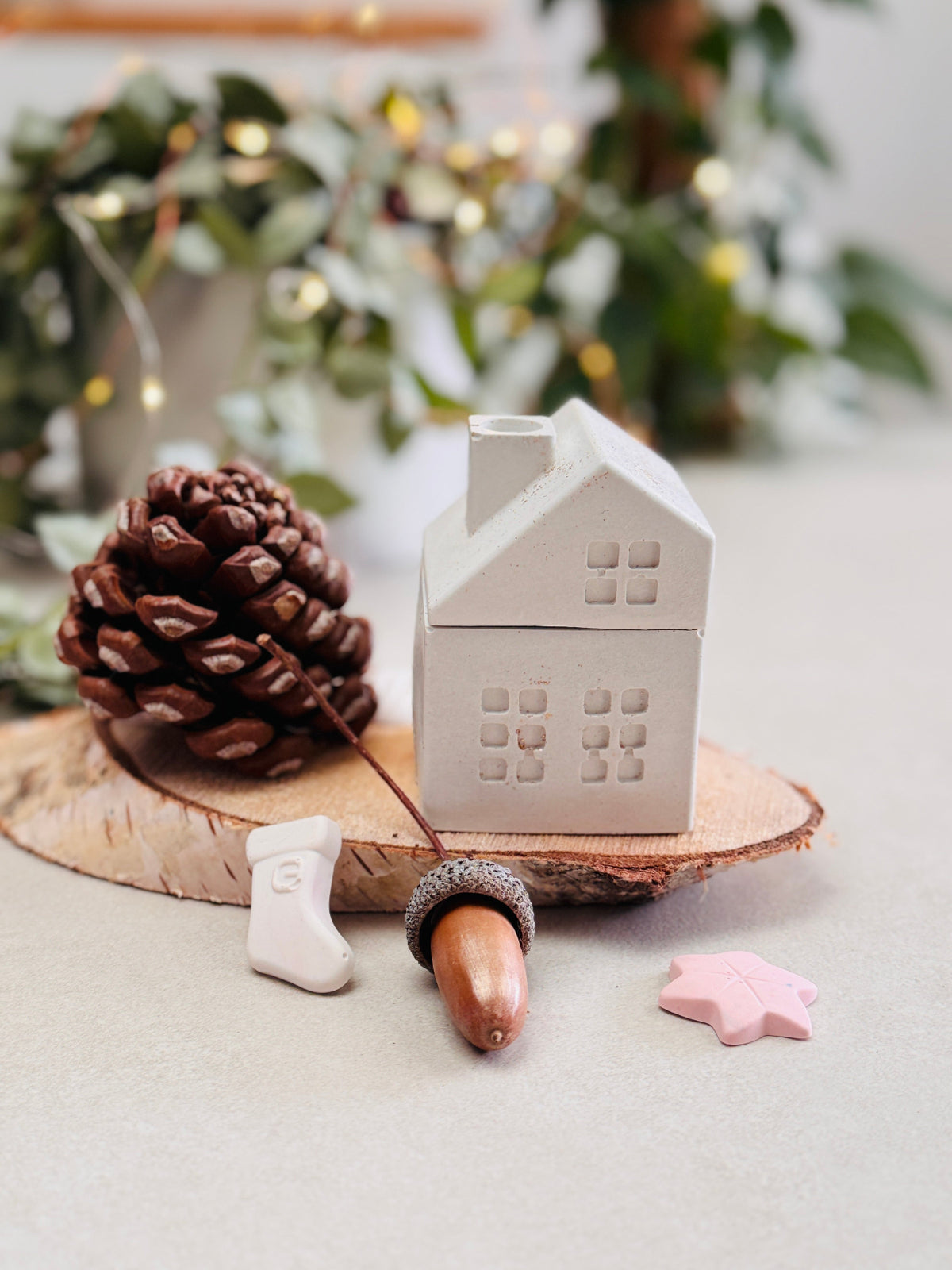 Bougie blanche en forme de maison avec cheminée posée sur un rondin de bois, entourée de petites décorations de Noël, de pommes de pin et de glands, chausson de père Noël.
