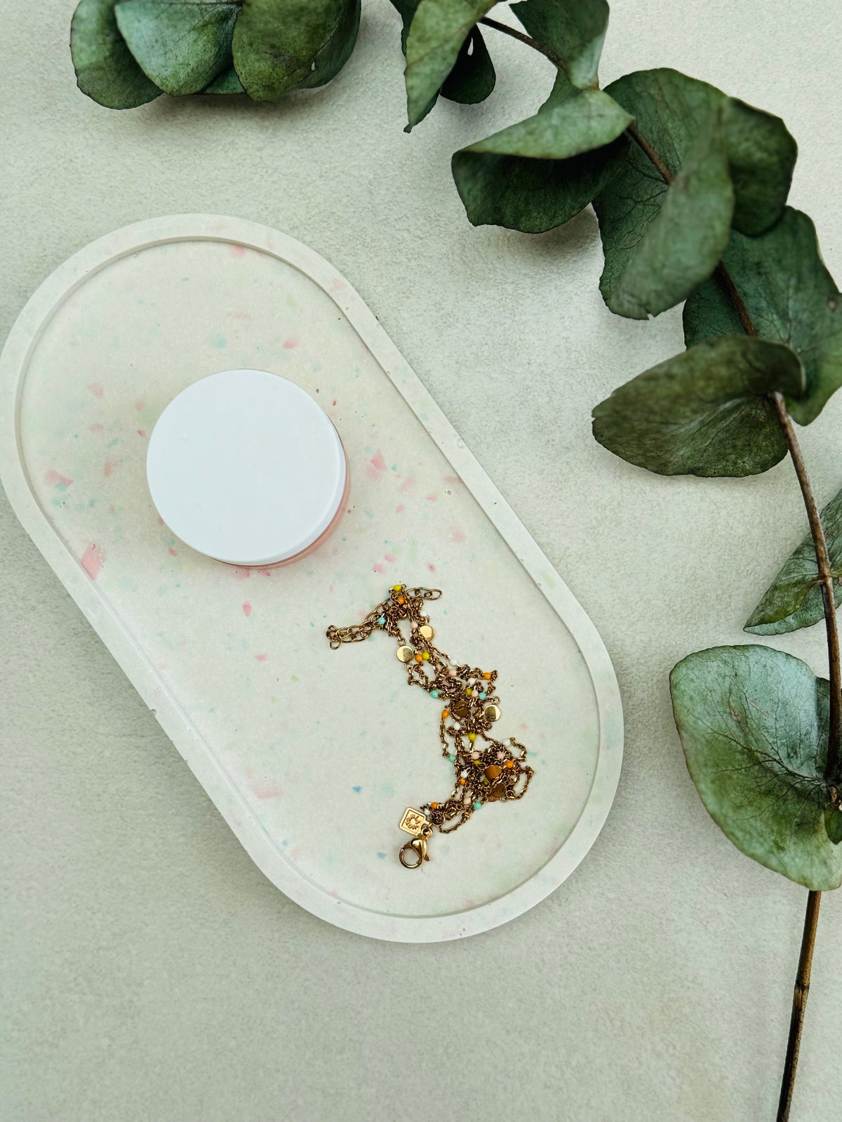 Plateau ovale en Jesmonite blanc avec des éclats de terrazzo rose et vert pastel, avec un bracelet doré et un  produit de soin.
