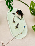 Plateau ovale en Jesmonite vert clair moucheté effet terrazzo, avec quelques bijoux et un produit de soin.