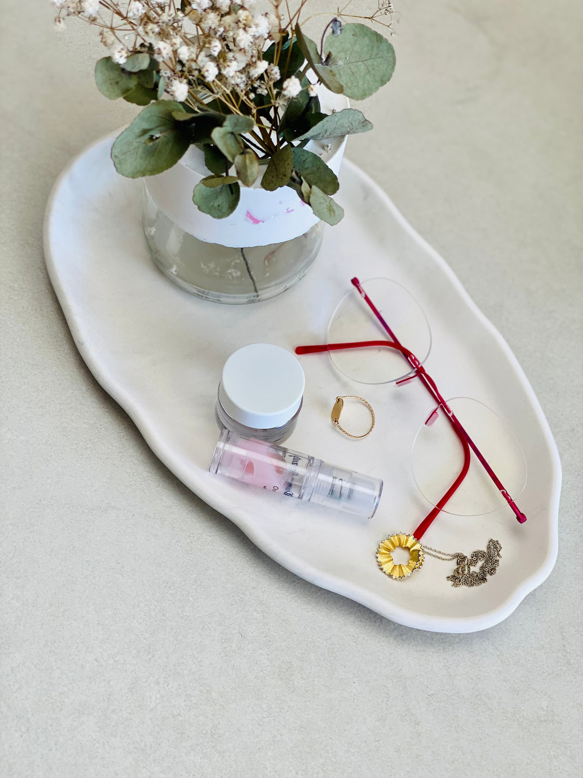Plateau ovale irrégulier blanc en Jesmonite avec bijoux, lunettes, soins et mini-vase fleuri