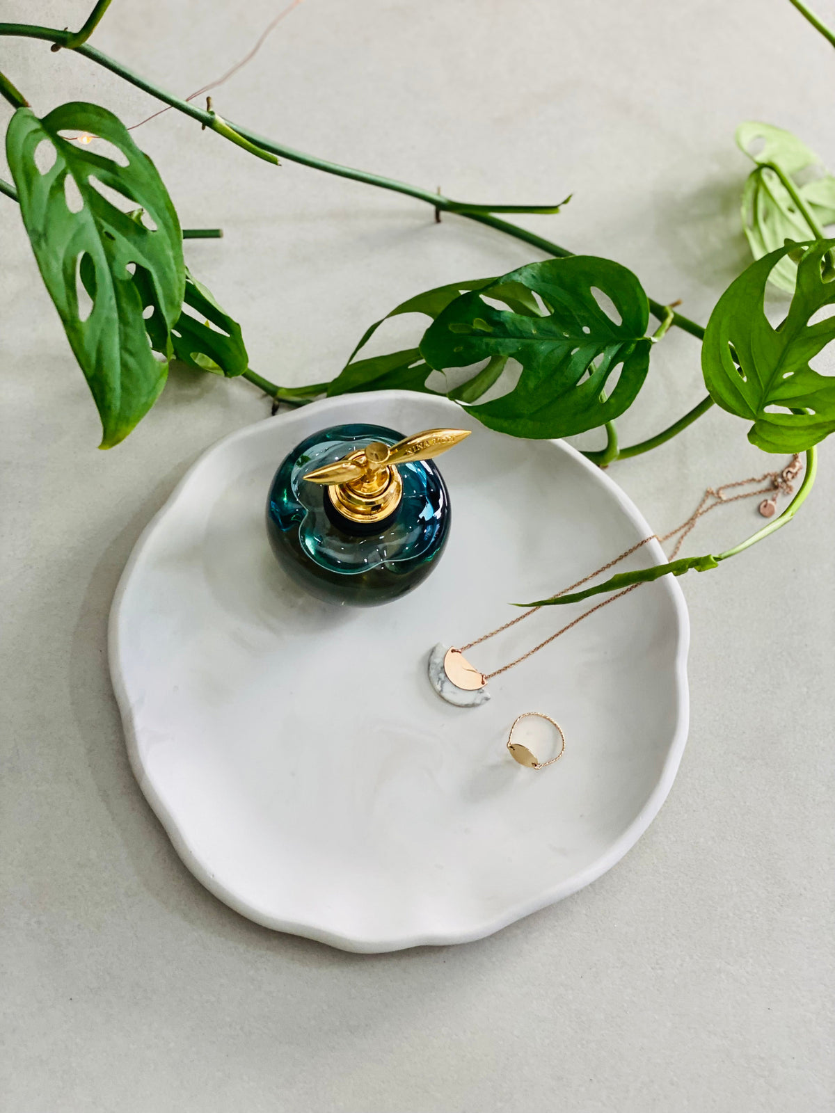 Plateau rond en Jesmonite blanc aux légers effets marbrés gris, avec un parfum et des bijoux, entouré de feuilles de plantes