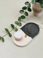 Plateau vide-poche ovale noir et blanc à effet terrazzo, avec des bijoux dorés et un petit pot à couvercle blanc.