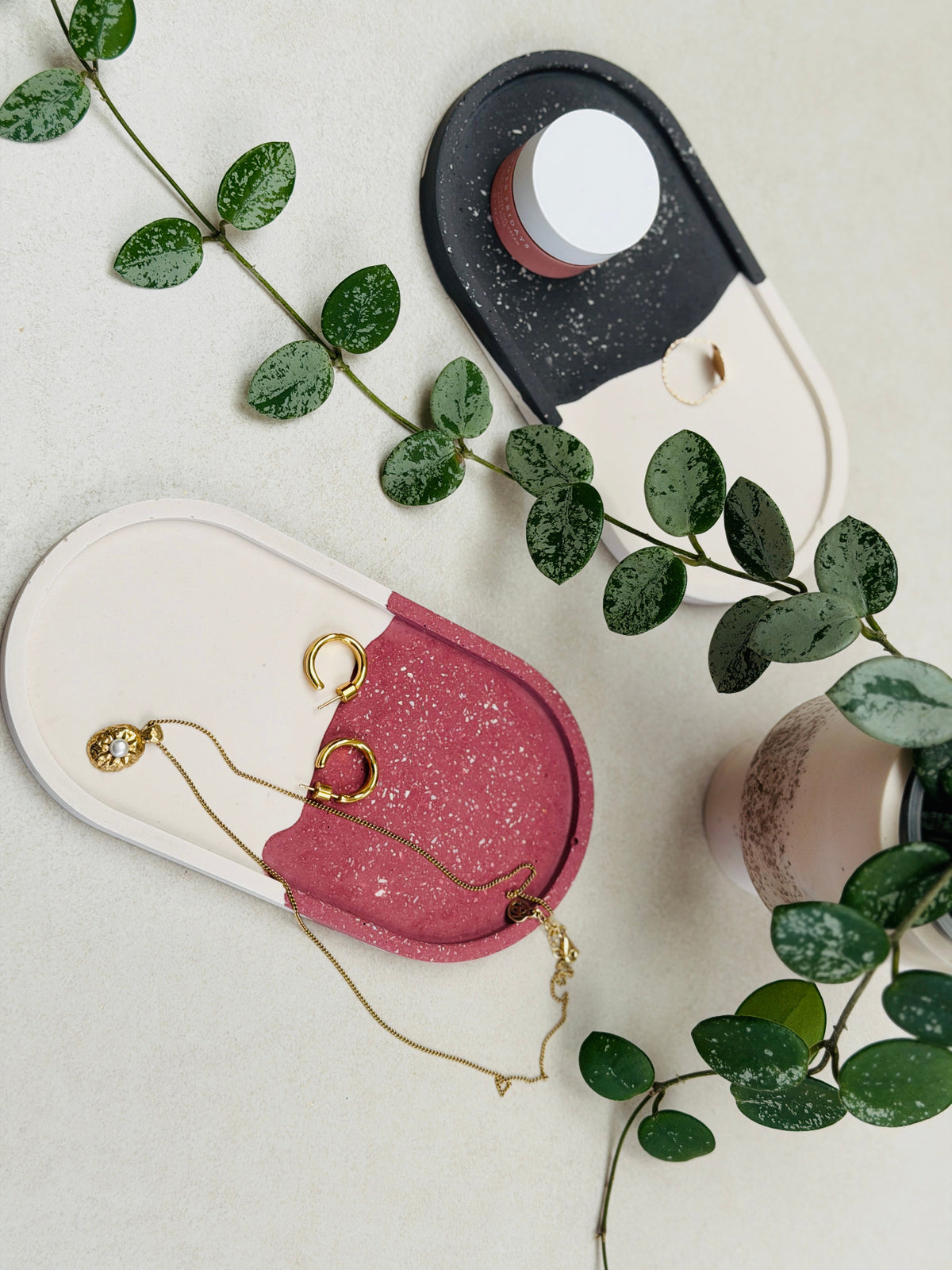 Plateaux jesmonite ovales à effet terrazzo bordeaux et noir sur fond blanc, utilisés pour ranger des bijoux et un petit pot de crème 