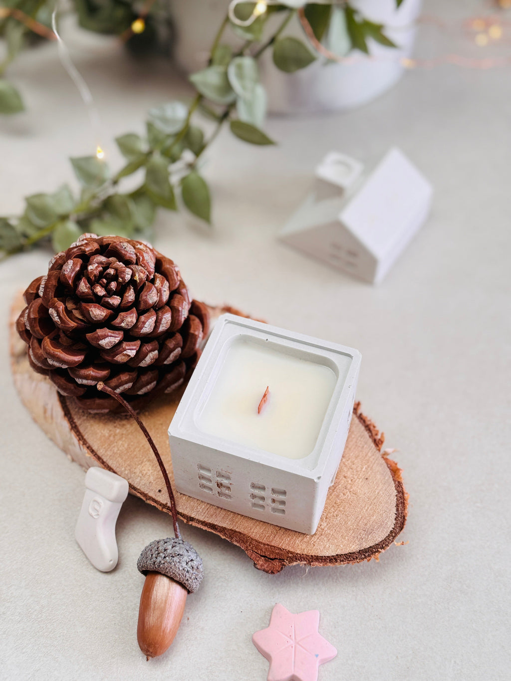 Bougie en forme de maison posée sur un rondin de bois, avec son couvercle en forme de toit et cheminée placé à côté, entourée de petites décorations de Noël, de pommes de pin et de glands.
