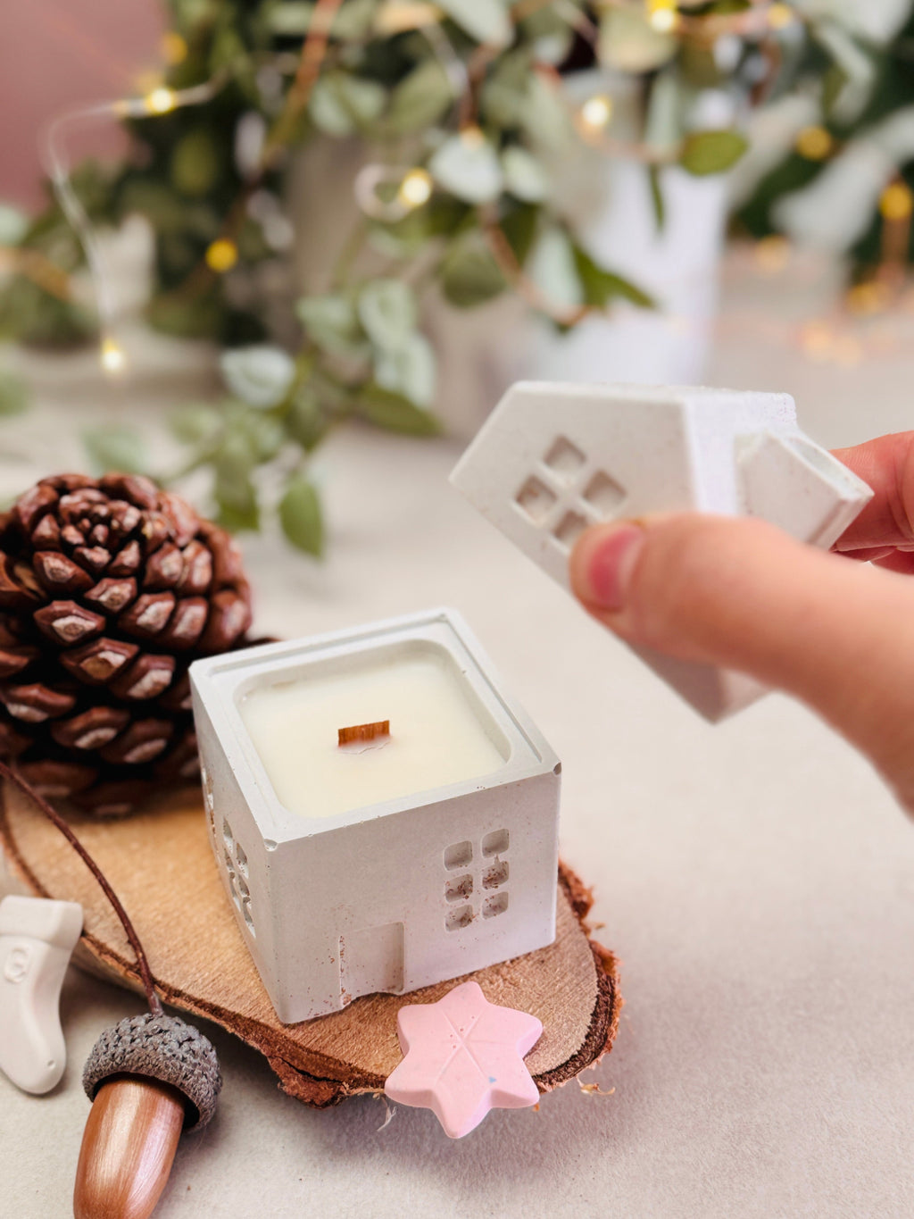 Couvercle de bougie en forme de toit avec cheminée, tenu dans une main au-dessus d’une bougie en forme de maison, entouré de petites décorations de Noël, de pomme de pin et de gland etc.

