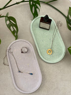Deux plateaux ovales en Jesmonite, l’un vert clair et l’autre lilas, avec un effet terrazzo, accompagnés de quelques bijoux et produits de soin.