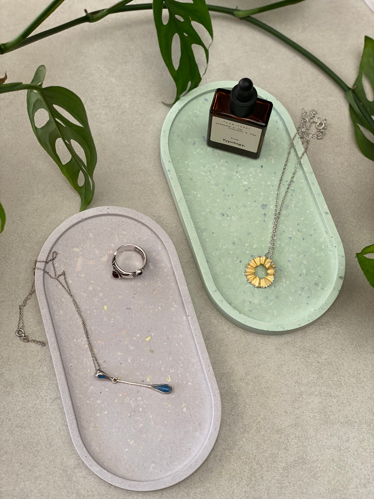 Deux plateaux ovales en Jesmonite, l’un vert clair et l’autre lilas, avec un effet terrazzo, accompagnés de quelques bijoux et produits de soin.