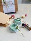 Kit DIY pour fabriquer des bougies florales, avec deux bougeoirs verts en béton, cire de soja, mèches en coton, fleurs séchées et accessoires de fabrication.
