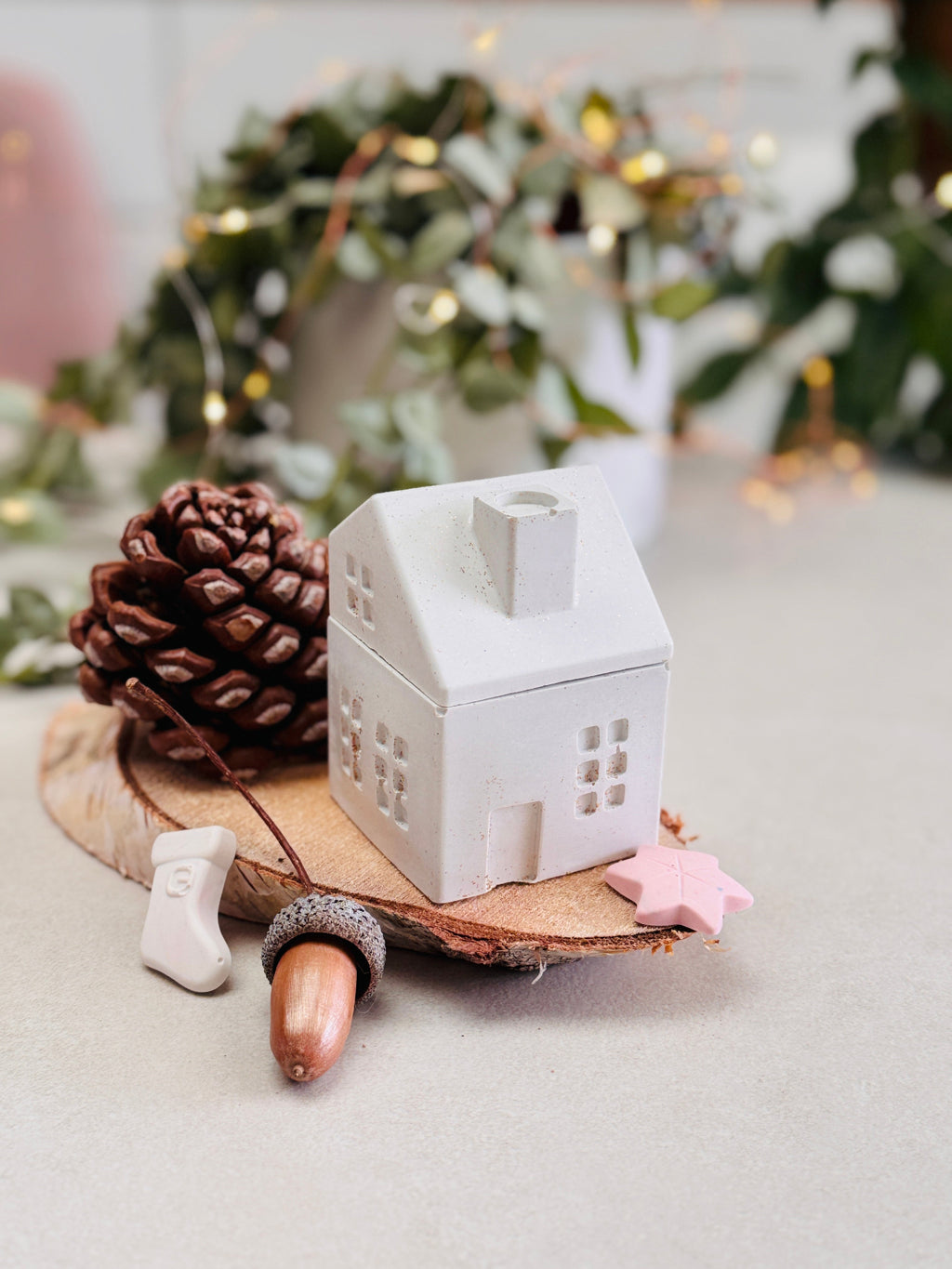 Bougie en forme de maison avec son couvercle en forme de toit et cheminée fermé, entourée de petites décorations de Noël, de pommes de pin et de glands, étoile. 
