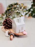 Bougie en forme de maison avec son couvercle en forme de toit et cheminée fermé, entourée de petites décorations de Noël, de pommes de pin et de glands, étoile. 

