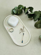 Présentoir ovale en Jesmonite blanc avec des éclats de terrazzo rose et vert pastel, quelques bijoux et une boîte d’écouteurs sans fil.
