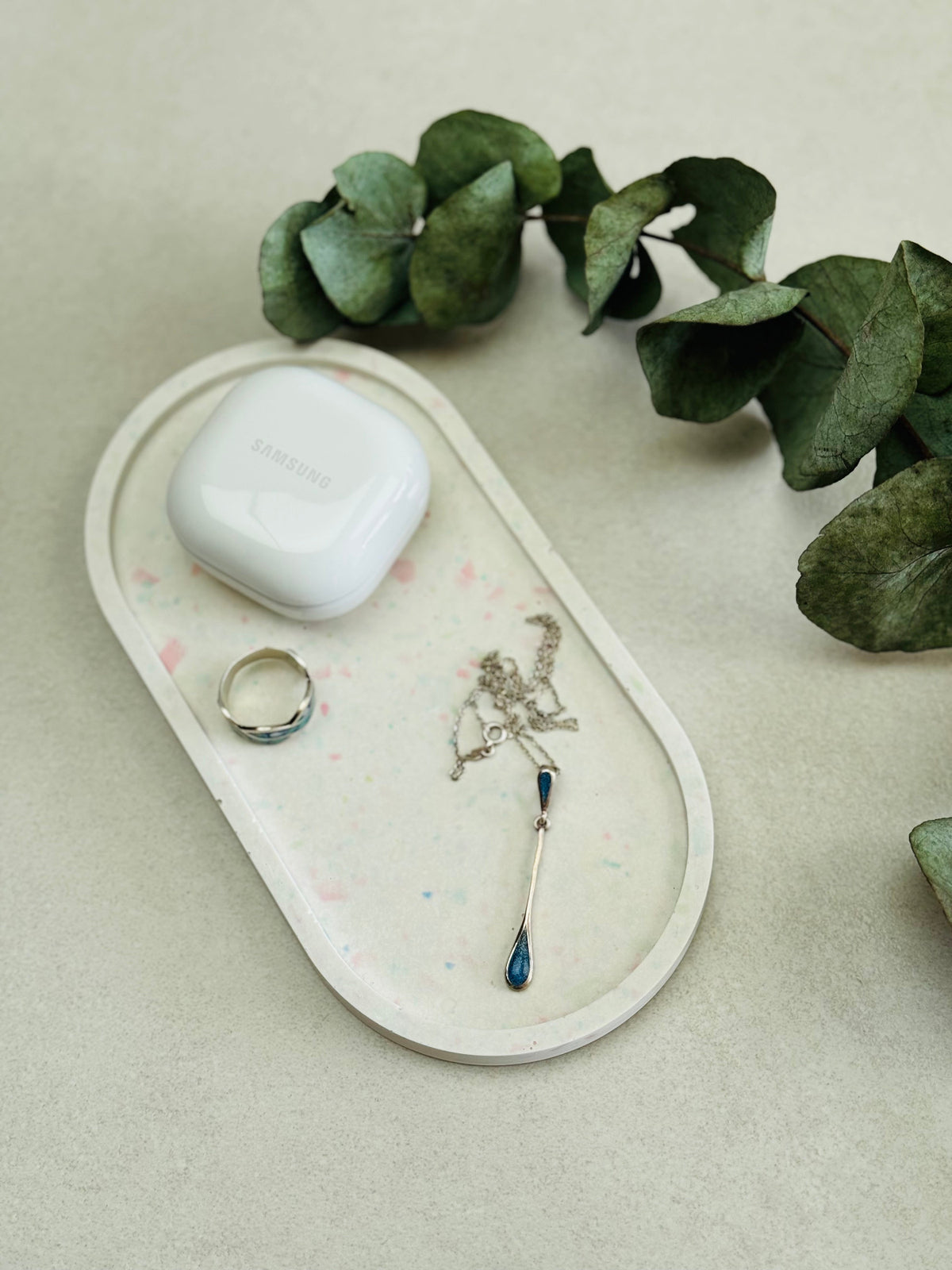 Présentoir ovale en Jesmonite blanc avec des éclats de terrazzo rose et vert pastel, quelques bijoux et une boîte d’écouteurs sans fil.
