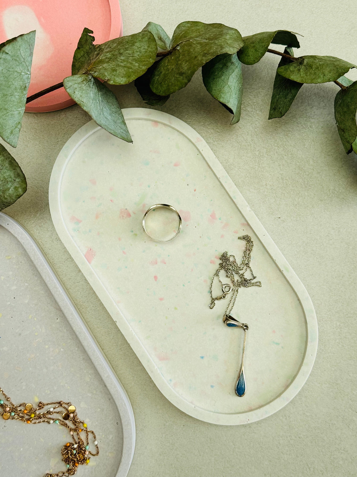Plateau ovale en Jesmonite blanc avec des éclats de terrazzo rose et vert pastel, avec quelques bijoux, tandis qu’un second plateau lilas apparaît partiellement.